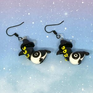 May include: A pair of handmade earrings featuring a whimsical design. Each earring showcases a black and white bird-like figure with yellow accents and a small black hat. The earrings have black ear wires and are set against a blue and purple background.