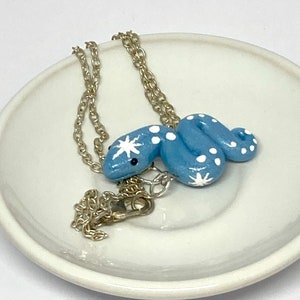 May include: A light blue snake pendant necklace with white polka dots and star accents. The snake pendant is on a silver chain and rests on a white ceramic dish. The necklace is a whimsical accessory.