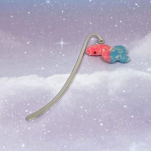 May include: A silver metal bookmark with a red and blue gummy bear charm. The bookmark has a curved design and is set against a background of a cloudy sky with stars.