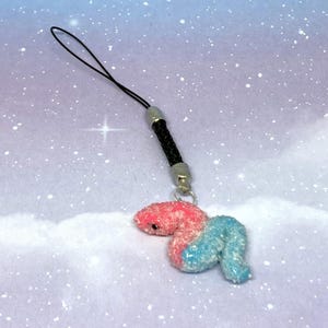 Puede incluir: Un pequeño amuleto hecho a mano con una criatura rosa y azul con forma de gusano. El amuleto está unido a un cordón negro con un cierre de metal plateado. El fondo es una suave y soñadora mezcla de azul y morado con motas blancas.