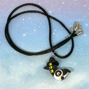 May include: A black cord necklace featuring a pendant shaped like a stylized bird. The pendant is black and white with yellow accents and a small black hat. The necklace has a silver-colored clasp.