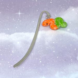 May include: A whimsical bookmark featuring a silver metal stem and a colorful worm charm. The worm is orange and green, with a textured, speckled appearance. The bookmark is designed for book lovers and makes a unique gift.
