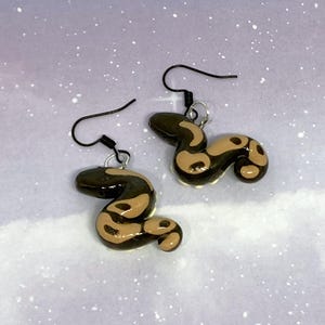 May include: A pair of handmade snake earrings. Each earring features a small, coiled snake design with a black head and tail, and a tan body with dark brown spots. The earrings have black ear wires and are set against a soft, speckled background.