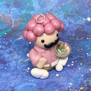 Puede incluir: Una figurita de arcilla rosa y blanca de una persona con un peinado rizado rosa que sostiene un vaso transparente con una aceituna verde dentro. La figurita lleva una camisa rosa y pantalones marrones.