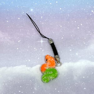 Puede incluir: Un colorido charm para teléfono con tres formas abstractas en naranja y verde, cubiertas con una sustancia blanca y brillante. El charm está unido a un cordón negro con un cierre de metal plateado.