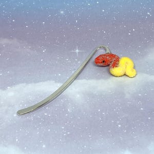 May include: A silver metal bookmark with a decorative red and yellow flower design. The flower has a textured, glittery appearance. The bookmark is curved and has a smooth finish. The background is a soft blue with a starry effect.