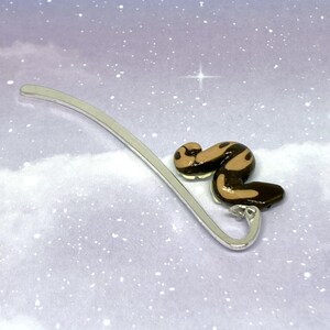 May include: A silver metal bookmark with a decorative snake design. The snake is brown and tan with a glossy finish. The bookmark has a curved top and a loop at the bottom.