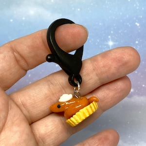 May include: A miniature pie charm with a black clip. The pie is orange with a white dollop of cream and sits in a yellow crust. The charm is attached to a black clasp.