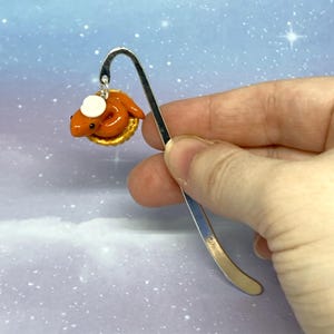 May include: A silver bookmark with a curved top. It has a small, orange, cartoon-like creature with a white hat, on a golden base. The background is a soft blue with white specks.