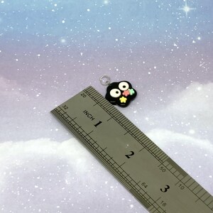 May include: A black charm with white eyes and colorful star accents. The charm is approximately 1 inch in size and has a silver loop for attaching to a necklace or bracelet. The background is a light blue and purple gradient.