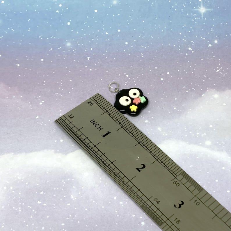 May include: A black charm with white eyes and colorful star accents. The charm is approximately 1 inch in size and has a silver loop for attaching to a necklace or bracelet. The background is a light blue and purple gradient.