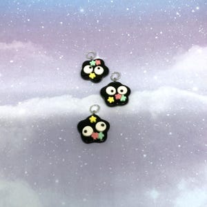 May include: Three black charms with white eyes and colorful star accents. Each charm has a silver loop at the top for attaching to a necklace or bracelet. The charms are set against a soft, pastel background.
