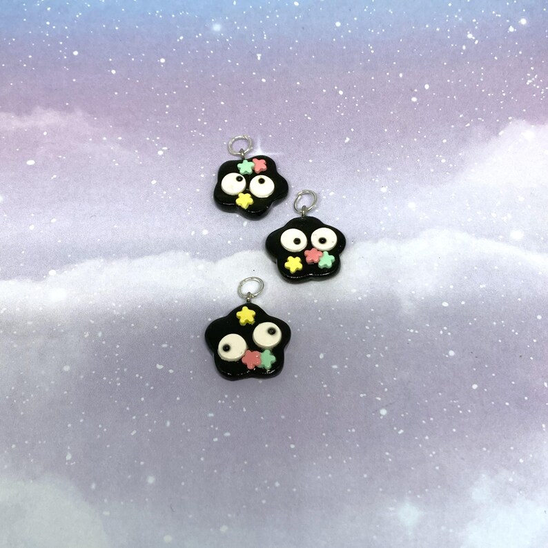 May include: Three black charms with white eyes and colorful star accents. Each charm has a silver loop at the top for attaching to a necklace or bracelet. The charms are set against a soft, pastel background.