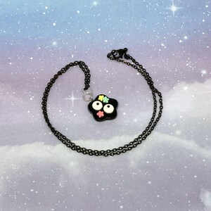 May include: A black necklace with a pendant shaped like a black flower with white eyes and colorful flower accents. The chain is black and the background is a cloudy sky with stars.