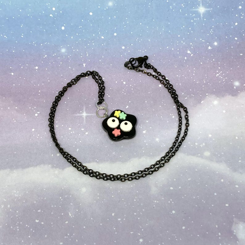 May include: A black necklace with a pendant shaped like a black flower with white eyes and colorful flower accents. The chain is black and the background is a cloudy sky with stars.