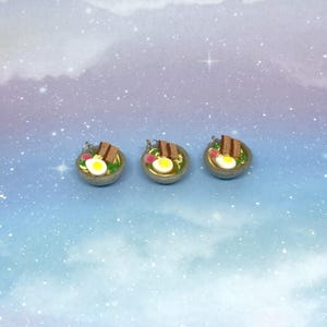 May include: Three miniature ramen bowls with detailed toppings, including a fried egg, meat, and vegetables. The bowls are gray with a gold rim, set against a blue and white background. These handcrafted food-themed charms are perfect for jewelry or craft projects.