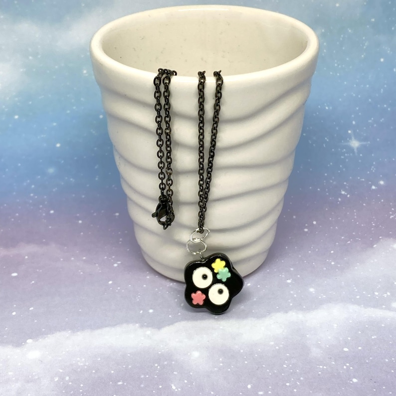 May include: A black necklace with a flower-shaped pendant. The pendant has a black base with white circles and colorful flower accents. The necklace chain is black and hangs inside a white ceramic vase.