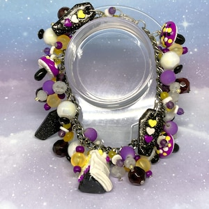 May include: A charm bracelet with a silver chain and various colorful beads. The charms include black coffin shapes with yellow and white hearts, purple mushroom shapes, and white and black shapes. The beads are purple, yellow, white, and clear.