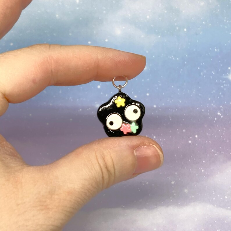 May include: A black star-shaped charm with white eyes and colorful star accents. The charm is held between fingers against a soft, blurred background. The charm has a silver loop for attaching to a necklace or bracelet.
