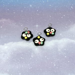 May include: Three black charm pendants with white eyes and colorful star accents. Each charm has a silver loop at the top for attaching to jewelry. The background is a light blue with a starry sky effect.