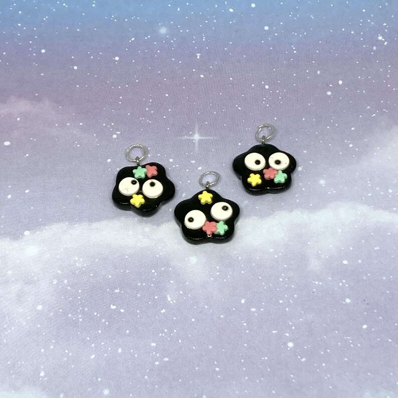 May include: Three black charm pendants with white eyes and colorful star accents. Each charm has a silver loop at the top for attaching to jewelry. The background is a light blue with a starry sky effect.