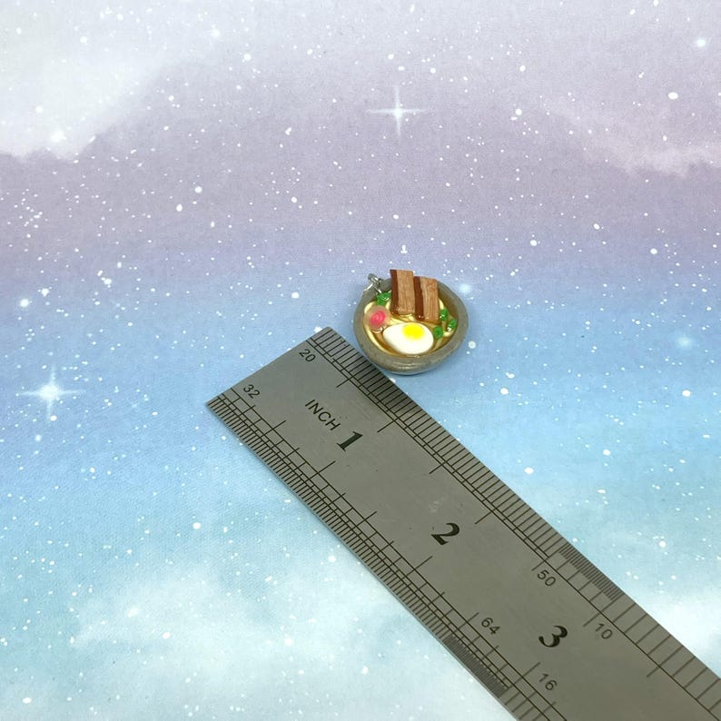 May include: Miniature ramen bowl charm with a fried egg, noodles, and meat. The charm is approximately 1 inch in diameter. The charm is on a blue and white background with a ruler.