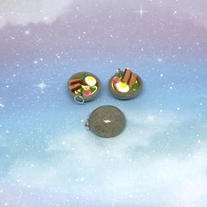 May include: Three miniature food charms. Two are bowls of ramen with egg, meat, and vegetables. The third is a gray, speckled, round charm. All three charms have a silver-colored loop for attaching to a necklace or bracelet.