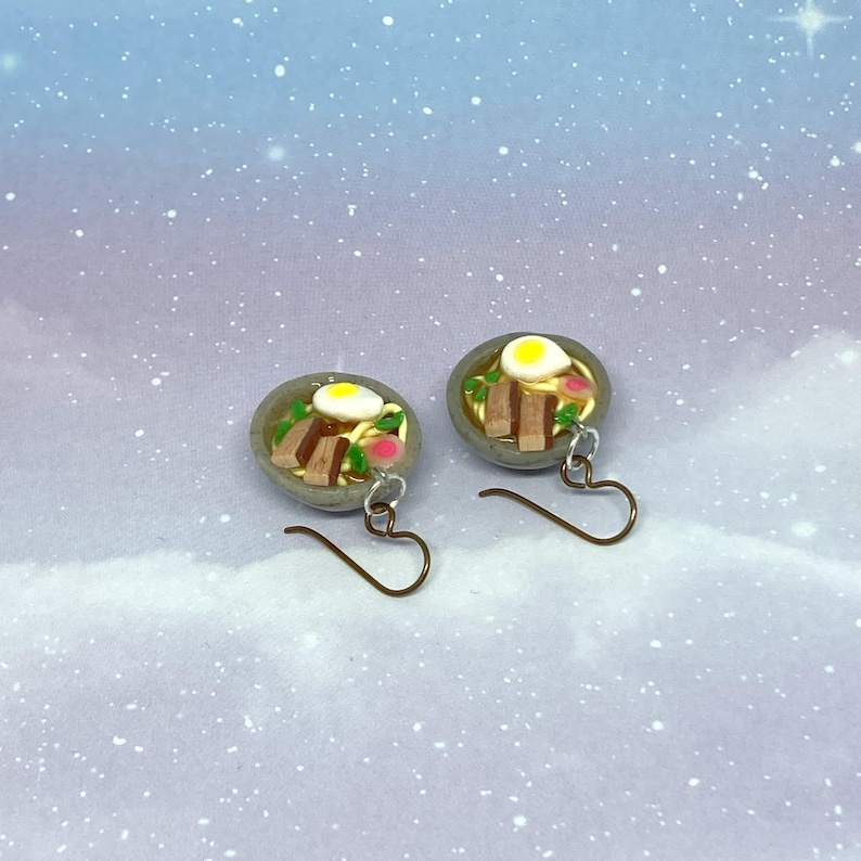 May include: A pair of miniature ramen bowl earrings. Each earring features a gray bowl filled with noodles, a fried egg, and other toppings. The earrings have bronze-colored hooks and are set against a blue and white speckled background.