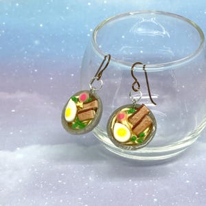 May include: A pair of handmade ramen bowl earrings. Each earring features a miniature bowl with a yellow broth, a half-boiled egg, pork belly slices, green scallions, and a pink swirl. The earrings hang from bronze-colored ear wires.