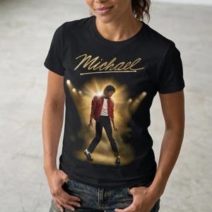 Może przedstawiać: Czarny t-shirt ze złotym napisem "Michael" i wizerunkiem artysty w czerwonej kurtce i czarnych spodniach. Osoba ma na sobie niebieskie dżinsy.