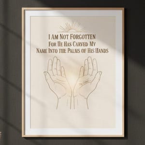 May include: Framed art print with a beige background and a minimalist design. The artwork features two open hands with the text "I AM NOT FORGOTTEN FOR HE HAS CARVED MY NAME INTO THE PALMS OF HIS HANDS" in brown. The frame is a light wood color.