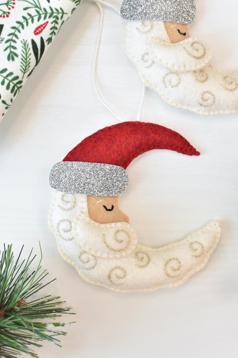 May include: Felt Christmas ornaments in the shape of a crescent moon wearing a Santa hat. The ornaments are white with gold stitching, a red hat, and a silver glitter band. Festive holiday decor.