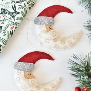 May include: Two crescent moon Christmas ornaments. Each ornament has a red felt hat with a silver glitter band, a tan face, and a white body with embroidered swirls. A sprig of evergreen and a patterned fabric are in the background.