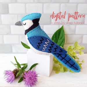 以下が含まれることがあります： 青い鳥のフェルトクラフトパターンとステップバイステップのチュートリアル。青い鳥は、緑の葉と紫の花を背景に、白いブロックの上に座っています。パターンには、白いお腹と黒い目をした青い鳥が含まれています。鳥は頭に黒いストライプがあり、尾は黒のアウトラインで青い色をしています。