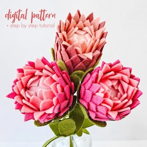 Op de afbeelding: Een boeket van drie roze protea bloemen van vilt met groene viltbladeren. De bloemen zijn in een vaas gerangschikt. De afbeelding bevat de tekst "digital pattern + step by step tutorial".