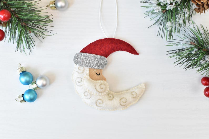 May include: A felt Christmas ornament shaped like a crescent moon with a Santa Claus face. The ornament has a red hat with a silver glitter band, a cream-colored face, and a white beard with embroidered swirls. The ornament hangs from a white string.