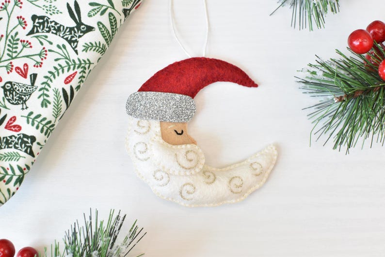 May include: A felt Christmas ornament shaped like a crescent moon wearing a Santa hat. The moon is white with embroidered details, a tan face, and a red hat with a silver glitter band. Green pine branches and red berries are in the background.