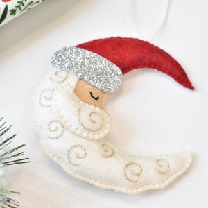 May include: A felt Christmas ornament shaped like a crescent moon wearing a Santa hat. The moon is white with gold embroidered swirls, a tan face, and a closed eye. The Santa hat is red with a silver glitter band.