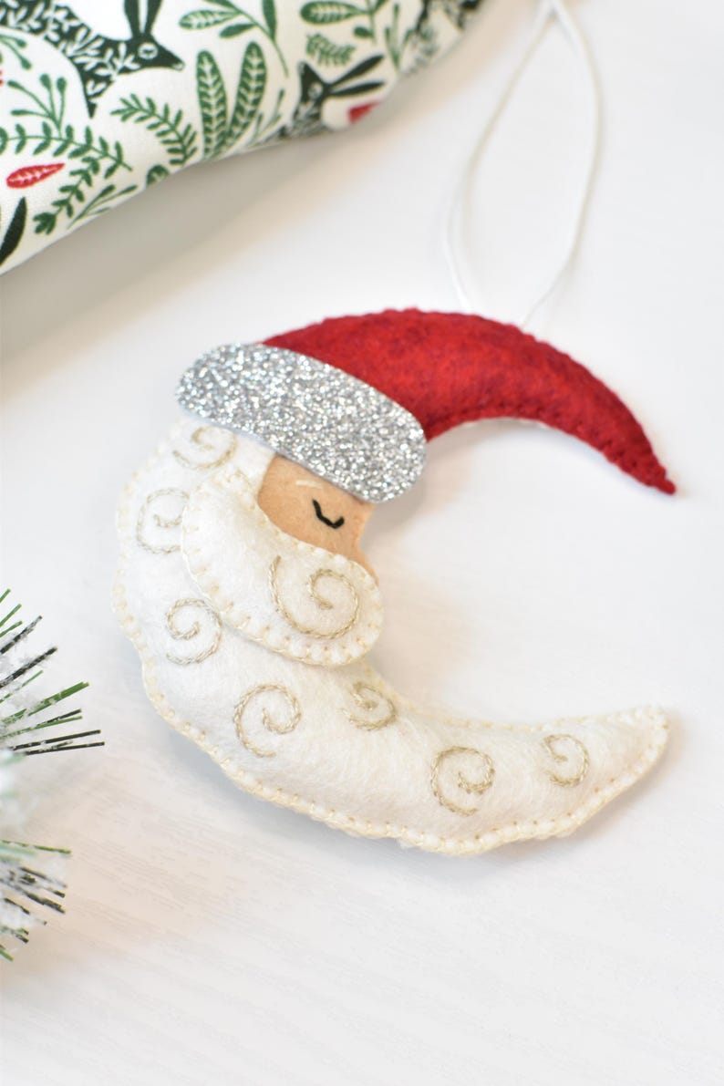 May include: A felt Christmas ornament shaped like a crescent moon wearing a Santa hat. The moon is white with gold embroidered swirls, a tan face, and a closed eye. The Santa hat is red with a silver glitter band.