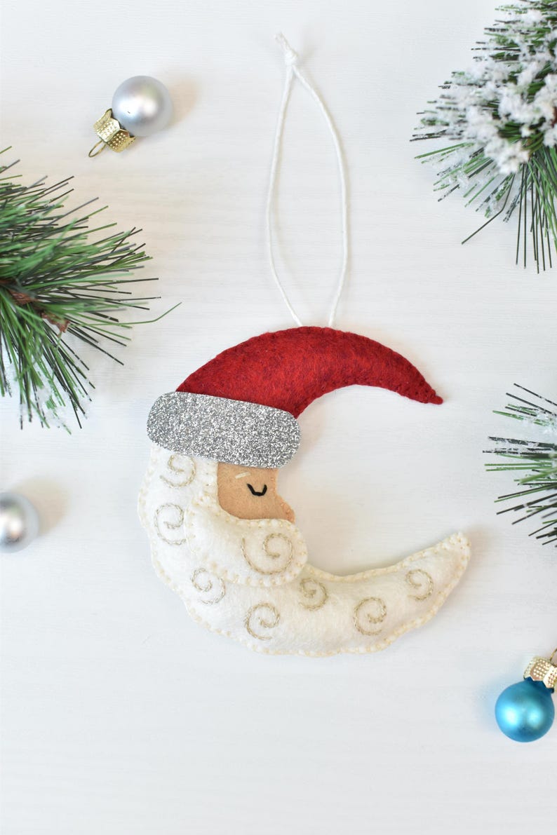 May include: A felt Christmas ornament shaped like a crescent moon with a Santa Claus face. The moon is off-white with gold stitching, a red hat, and a silver glitter brim. A white string is attached for hanging. Christmas decor.