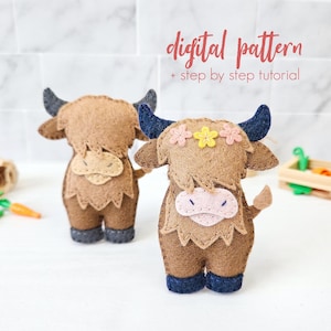Peut inclure: Deux peluches de vache écossaise en feutre, l'une avec un corps brun et l'autre avec un corps brun et une couronne de fleurs. Les peluches ont des yeux noirs et des nez roses. Le texte "digital pattern + step by step tutorial" est écrit sur un fond blanc.