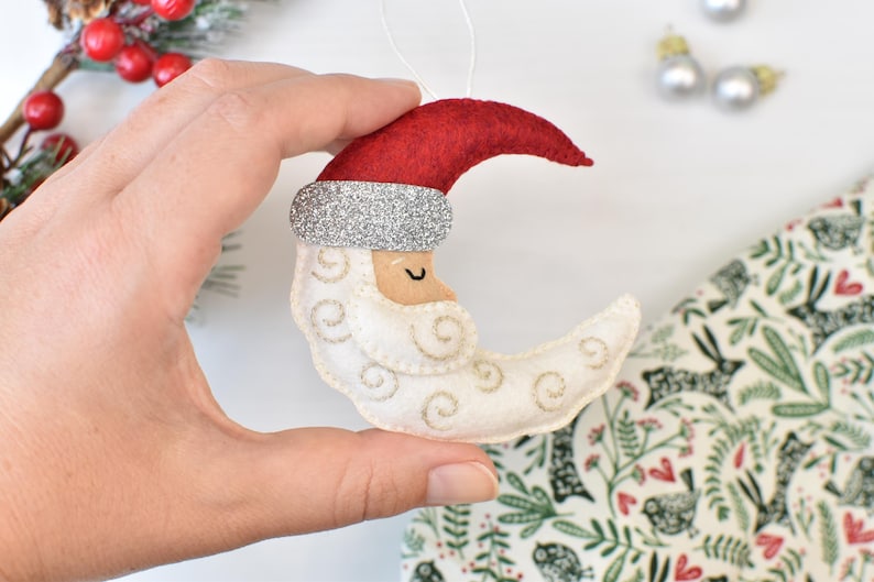 May include: A felt Christmas ornament shaped like Santa Claus in a crescent moon. The ornament features a red hat with a silver glitter band, a cream-colored face and beard, and gold embroidered details. A white string is attached for hanging.