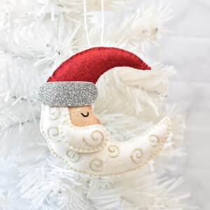 May include: A felt Christmas ornament shaped like a crescent moon with a Santa Claus face. The ornament features a red hat, silver glitter trim, and embroidered details. It hangs from a white string.