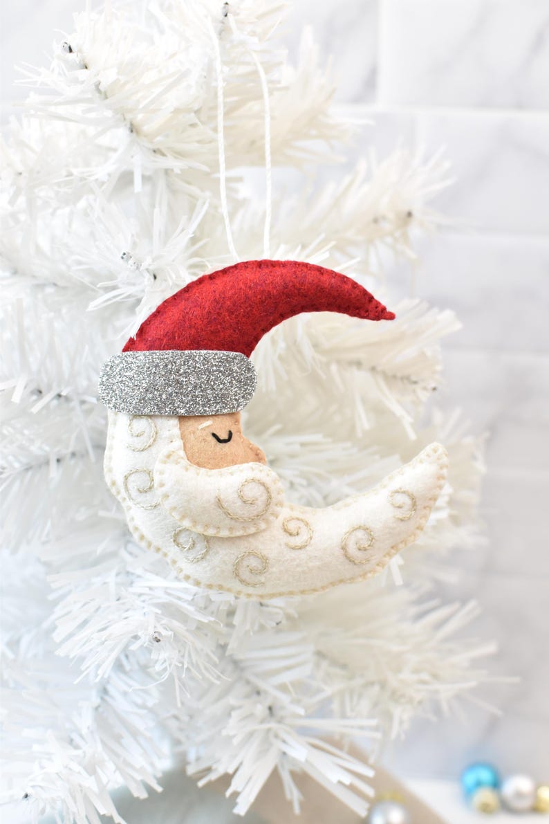 May include: A felt Christmas ornament shaped like a crescent moon with a Santa Claus face. The ornament features a red hat, silver glitter trim, and embroidered details. It hangs from a white string.