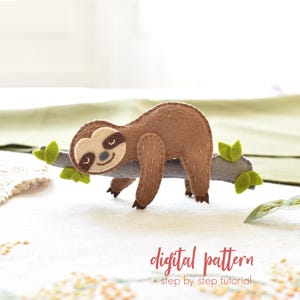 May include: A brown felt sloth figurine resting on a gray felt branch with small green leaves. The sloth has a cream-colored face with dark brown eye patches and stitched details. The text digital pattern + step by step tutorial is visible.
