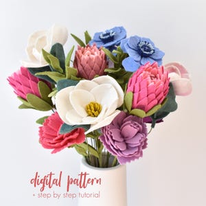Puede incluir: Un ramo de flores de fieltro en un jarrón blanco. El arreglo incluye flores blancas, rosas, rojas, moradas y azules con hojas verdes. El texto "digital pattern + step by step tutorial" está en la parte inferior.