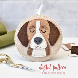 May include: A circular felt dog ornament with a beige base, brown ears, and a white and brown face. The dog has closed eyes and a black nose. It has a white ribbon for hanging. Text on the image reads "digital pattern + step by step tutorial."