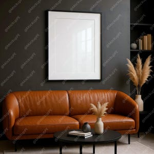 May include: A modern living room scene featuring a brown leather sofa, a black coffee table, and a framed blank picture on the wall. Decorative vases with dried plants and books are also visible. The room has a dark, neutral color palette.