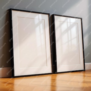 Pode incluir: Duas impressões de arte emolduradas em preto com passe-partouts brancos encostadas a uma parede cinza em um chão de madeira. As molduras são retangulares e têm um design simples e moderno. As impressões estão em branco.