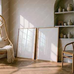 May include: Two large, blank picture frames with light wood frames lean against a wall in a well-lit room. The frames are positioned on a herringbone wood floor. A wicker chair and a shelf with decorative vases are also visible.